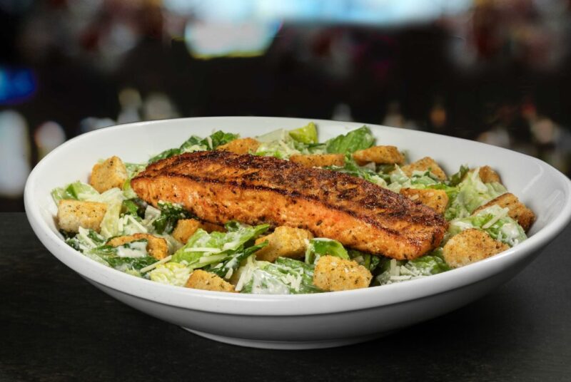 a plate of blackened salmon placed on top of a caesar salad